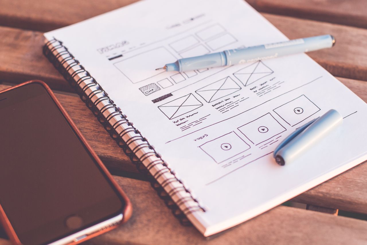 services-02 Vue rapprochée d’un carnet avec des esquisses de wireframe et un smartphone posé sur un bureau en bois.